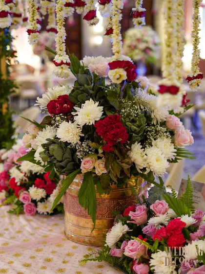 A lush and vibrant floral arrangement in a brass pot, featuring a mix of red, white, and pink flowers. The hanging floral strings (kaleeras) in the background add a traditional wedding touch.