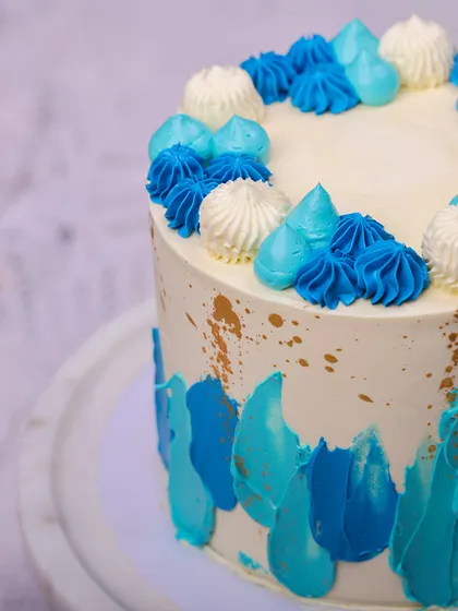 We create a piece of modern art with this abstract blue buttercream cake. Strokes of various blue shades are painted onto a white canvas, accented with gold splatters and topped with piped rosettes, making it a stylish and happy choice for a Father's Day celebration.