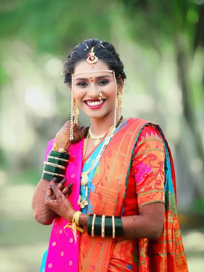 A happy portrait of the bride, her hand gently touching the mundavalya on her forehead. Her bright smile and traditional jewelry shine in this lovely outdoor shot.