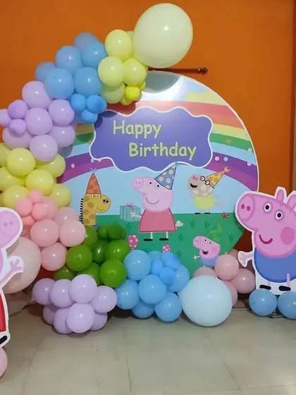A playful Peppa Pig themed birthday decoration. This setup includes cutouts of the entire Peppa Pig family against a custom backdrop, framed by a pastel-colored balloon arch.