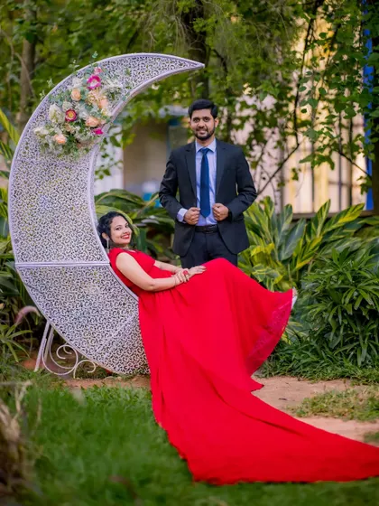 A relaxed and happy pre-wedding photo. The client is wearing a red flying gown with a long trail, seated on a crescent moon prop, while her partner stands by in a classic suit.