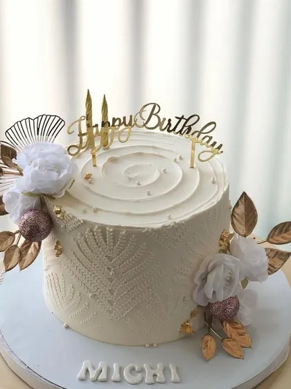 An elegant all-white cake with delicate lace-like stenciling and white and gold floral decorations.