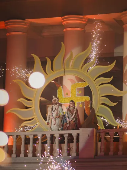 A picture-perfect varmala moment. The couple stands framed by a massive, custom-designed sun prop with a swastika, illuminated by sparklers. We design these central set pieces to create a stunning focal point for your ceremony.