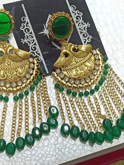 These antique gold finish earrings feature a peacock design with green stones and hanging chains. They are a perfect example of traditional temple jewellery design, ideal for wearing with sarees.