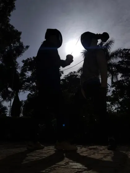 The moment before a bout begins, two fencers face each other, their concentration captured in this striking silhouette against the sun.