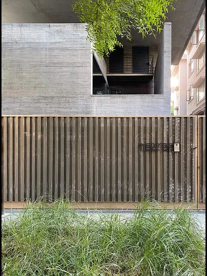 The entrance gate of the Jenga House, where a slatted wooden screen provides privacy while complementing the raw concrete structure above. The design integrates landscape elements at the threshold.