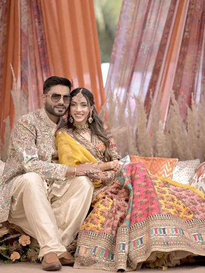 A beautiful portrait of the couple seated at their Mehndi, showcasing their stunning, colorful outfits against the rustic, draped backdrop.