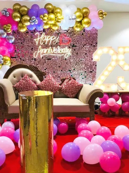 A 25th-anniversary party with a glamorous pink, purple, and gold theme. The setup includes a sequin wall, a comfortable sofa for photos, and marquee numbers.