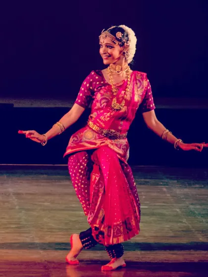 A vibrant stage photograph from a solo Bharatanatyam recital. The bright pink and gold costume, combined with the expressive smile, captures the joyful spirit of the dance.