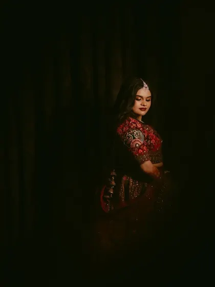 A moody and intimate portrait of a bride in a dark room. The soft lighting highlights her features and the rich details of her red lehenga.