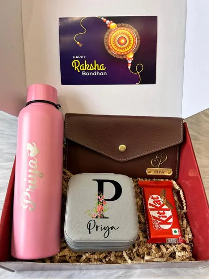 A pretty in pink Rakhi hamper for a sister. This gift set includes a personalized water bottle, a custom initial jewelry box, and a clutch, along with a KitKat.