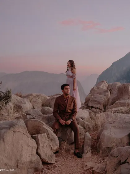 An editorial-style couple portrait set against a dramatic mountain landscape at dusk. The soft pink clouds and their poised expressions create a moody, romantic, and high-fashion feel.