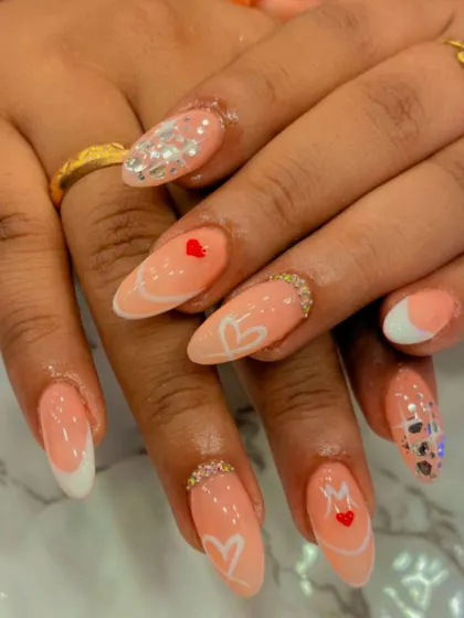 A personalized and romantic set of peach-colored almond nails. The design includes hand-painted hearts, initials, and delicate crystal embellishments.