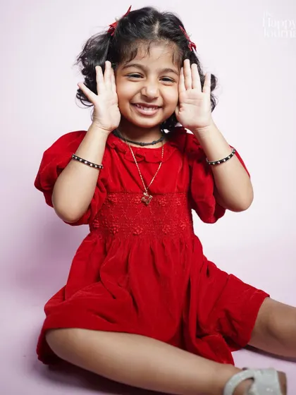 A little girl in a red dress plays peekaboo with a huge smile. Her joyful and playful nature shines through in this studio shot.