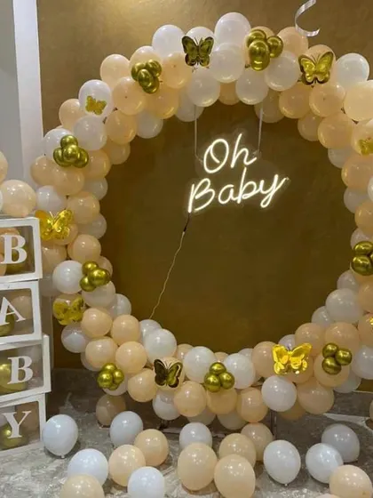 An elegant "Oh Baby" baby shower decoration. The setup features a full balloon ring in peach and white, with gold butterfly accents and "BABY" blocks.