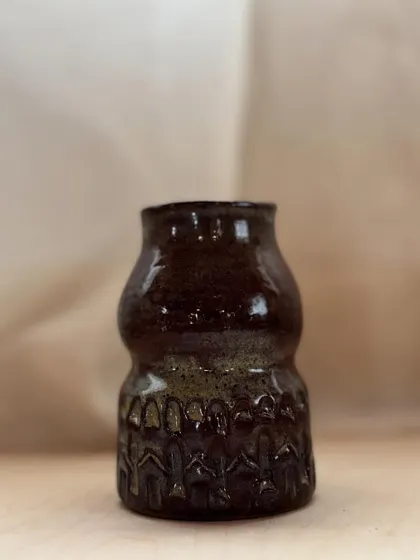 This dark, textured vase with intricate carvings was made by a student. The glossy finish highlights the detailed patterns, creating a piece that is both rustic and elegant.