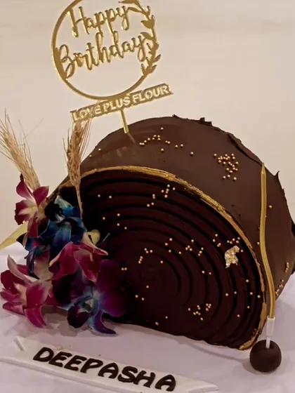 A unique and artistic chocolate cake for a birthday celebration. This log-shaped design is decorated with beautiful orchids and a custom name plaque, enjoyed here by a happy client.