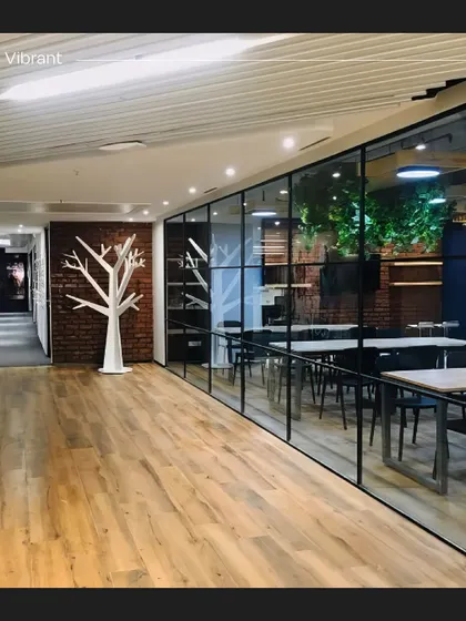 A wide shot of a hallway in the Crocs office, leading to a glass-enclosed meeting room. The design combines industrial elements like exposed brick with natural wood floors and a whimsical white tree sculpture, reflecting the brand's unique style.