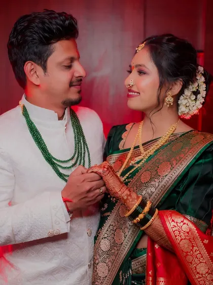 The couple holds hands and shares a loving gaze. This portrait highlights their traditional outfits, with the groom's emerald green necklace complementing the bride's beautiful saree.