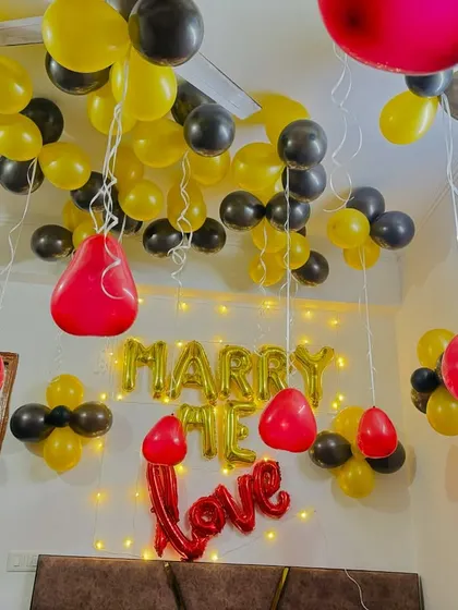 A 'Marry Me' proposal setup in a room filled with yellow and black balloons, with fairy lights adding a warm glow.