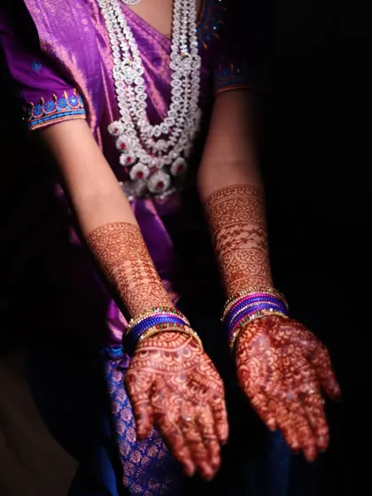A detailed shot focusing on the bride's intricate henna design. The dramatic lighting highlights the rich color of the mehndi against her purple and blue outfit.