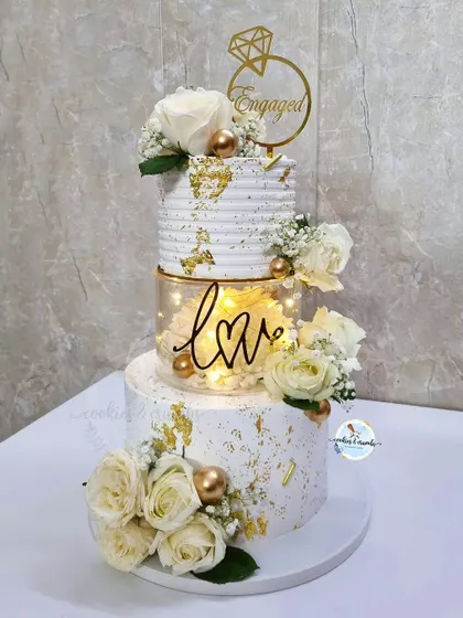 I love the drama of a lit-up tier. This three-tier engagement cake features a 'love' light, fresh white roses, and gold leaf accents, creating a stunning focal point for the celebration.