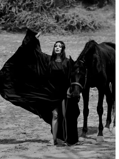 A full-length black and white shot of a model with a flowing cape standing next to a black horse, creating a majestic and powerful image.