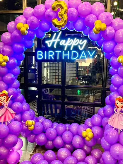 A magical princess-themed party for a 3rd birthday. This setup includes a full ring of purple balloons, a "Happy Birthday" neon sign, and cutouts of Princess Sofia.