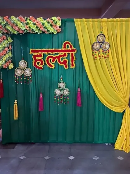 A simple yet elegant Haldi backdrop with a dark green curtain, a draped yellow accent, and traditional hanging tassels.
