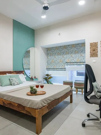 A bright and airy guest bedroom featuring a wooden bed, a teal accent wall, and a large round mirror. The room also includes a functional workspace, making it a versatile retreat for visitors.