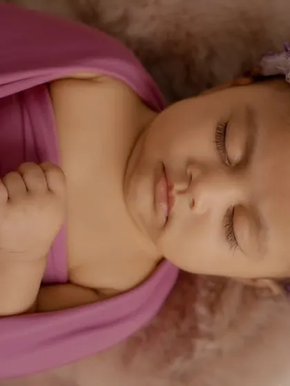 A close-up of a sleeping newborn wearing a purple floral headband, wrapped in a matching purple cloth.