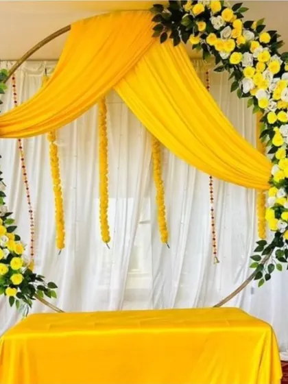 A clean and modern circular backdrop for a Haldi ceremony. The design uses yellow drapes and a half-arch of yellow and white flowers against a simple white curtain.
