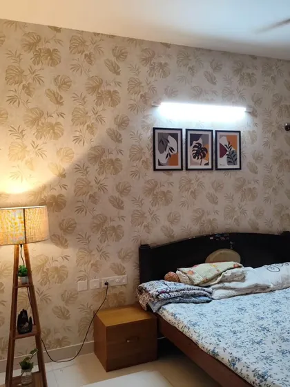A bedroom featuring a subtle monstera leaf wallpaper in a beige tone. The addition of a floor lamp and framed art shows how to style a wallpapered wall.