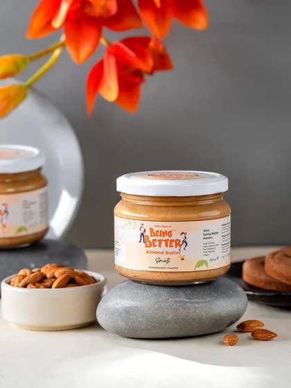 An elegant, minimalist shot of Being Better's almond butter jars resting on smooth stones. The addition of a single orange flower adds a touch of sophistication and natural beauty to the composition.