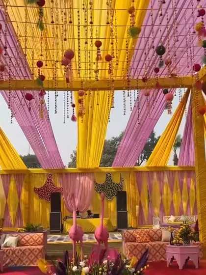 A festive Haldi event space with yellow and pink drapes, hanging tassels, and quirky flamingo props. This decor creates a fun and playful atmosphere for guests.
