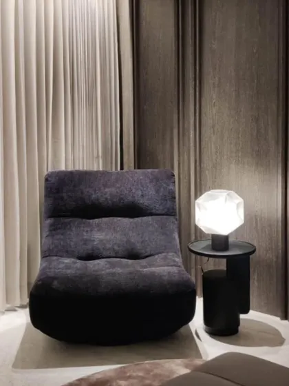 This cozy reading corner features a plush, low-slung lounge chair and a sculptural side table with a geometric lamp. The dark wood paneling and heavy curtains create a sense of enclosure and comfort.