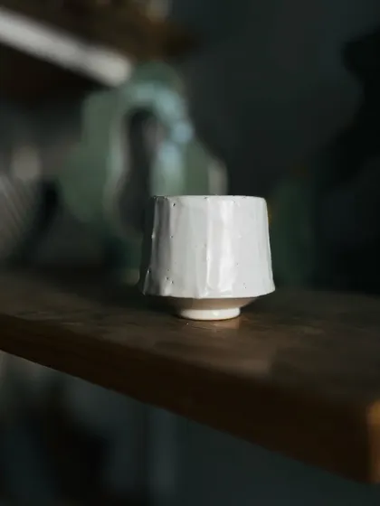 A small, faceted cup with a simple white glaze. The beauty is in the subtle, hand-carved shape that feels perfect to hold.