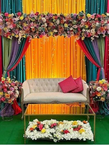 A rich and colorful Mehndi stage with a multi-hued drape backdrop. The vibrant floral arrangements and plush seating create a luxurious and festive atmosphere for the bride-to-be.