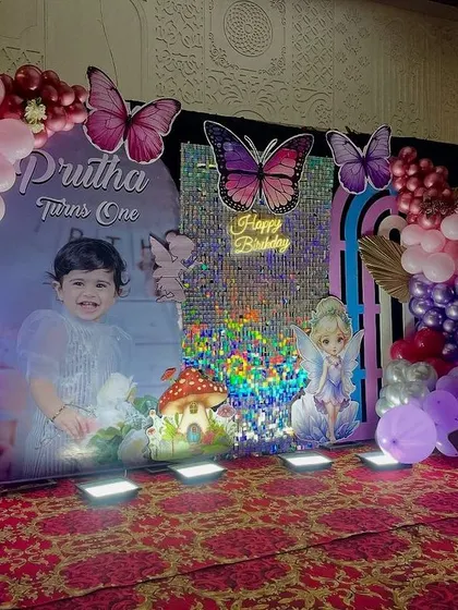 A butterfly-themed first birthday stage. The decor features a custom backdrop with the child's photo, a shimmering panel, and large butterfly cutouts, all framed by a pink and purple balloon arch.