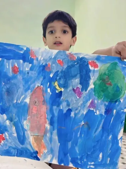 A young boy proudly displays his space-themed artwork, complete with a rocket ship, created in one of our online classes.