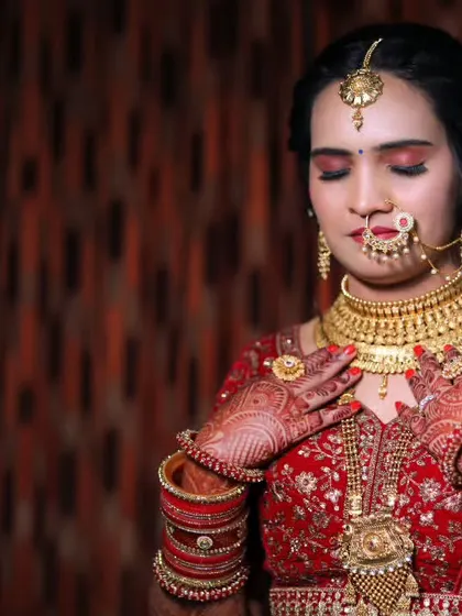 A stunning portrait of the bride in her red and gold wedding attire. She is adjusting her elaborate gold necklace, her eyes closed in a moment of quiet reflection before the ceremony.