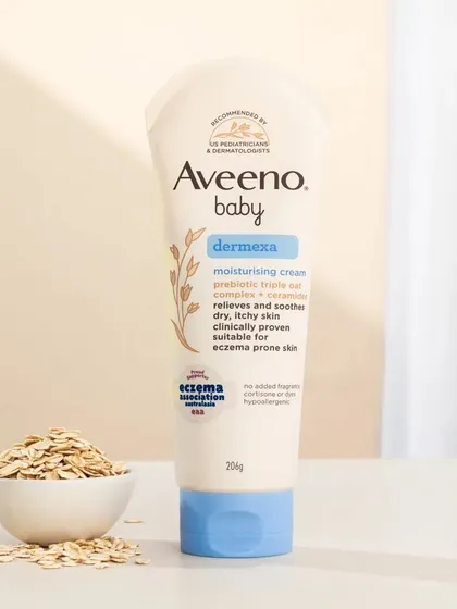 A hero shot of Aveeno Baby's Dermexa cream. The soft lighting and simple props like scattered oats create a gentle and caring mood, perfect for a baby care product.