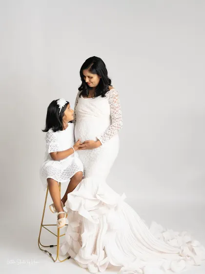 A touching portrait of a mother and her daughter. The little girl, seated on a stool, looks up at her mother, both dressed in elegant white lace outfits.