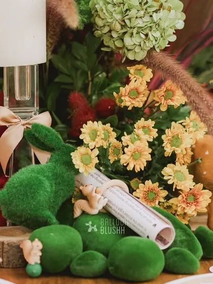 A close-up of a table centerpiece, featuring a mossy rabbit, a deer, and a scroll, all nestled among flowers.