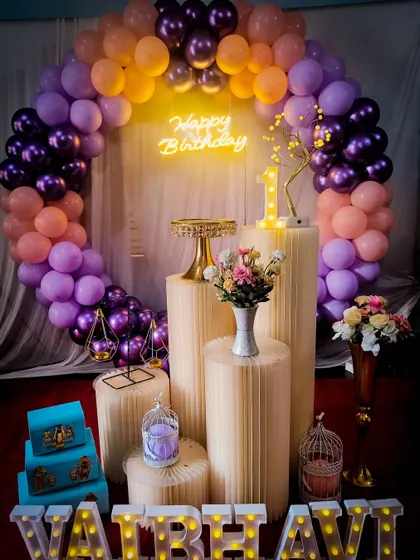 A beautiful first birthday setup with a purple and peach balloon arch and a 'Happy Birthday' neon sign.