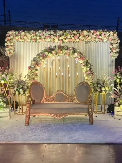 An enchanting outdoor evening setup for an engagement or Roka. A vintage sofa is placed under a floral arch, with candle-like lights and surrounding flower arrangements creating a magical ambiance.