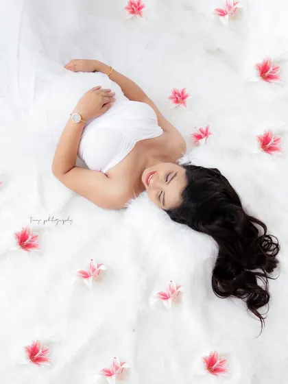 A creative and artistic overhead shot. The mom-to-be lies on a fluffy white rug, surrounded by scattered pink flowers, creating a beautiful and dreamy scene.