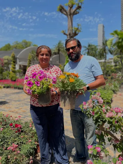 A happy customer couple holding their new flowering plants, surrounded by the beautiful blooms at our nursery.