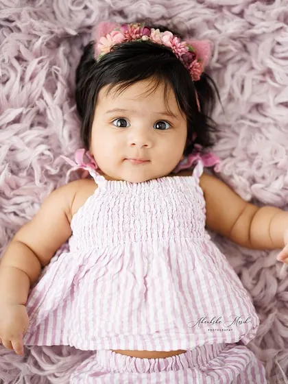 This sweet baby girl is lying on a soft, textured purple blanket, looking right at the camera. The floral cat-ear headband adds a touch of whimsy and fun.