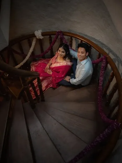 A high-angle shot of a couple on a dark wood spiral staircase. The composition draws the eye down, creating a sense of intimacy and romance within the architectural space.